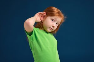 Portrait of cute redhead little girl trying to hear someone putting her hand on the ear, blue background, green t-shirt
